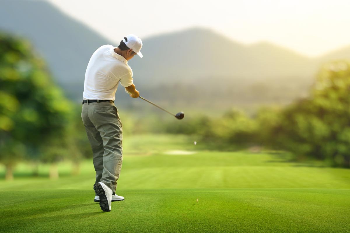 A golfer in a white shirt and cap mid-swing on a sunlit fairway, with hazy hills behind